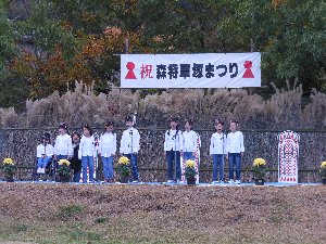 祝・森将軍塚まつりと書かれた幕の下で歌うジャージ姿の学生たちの写真