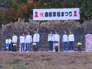 ジャージ姿の学生たちが、祝・森将軍塚まつりと書かれた幕の下で歌っている写真