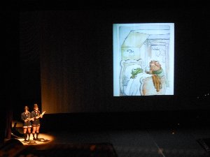 暗いステージの上で絵本の挿絵とともに英語の発表をする高校生女子二人の写真