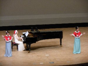 サンタ帽子を被ったピアノ奏者とケープを羽織った歌手二人がステージから手を振っている写真