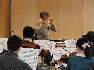 リハーサル中の指揮者の男性とバイオリン奏者4名が映った写真