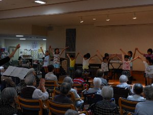 介護老人保健施設で歌を歌い、手を大きく振り上げる子供たちの写真