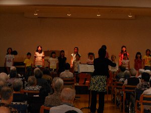 介護老人保健施設で歌を歌う子供たちとそれを聞く施設入居者の写真