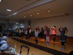 介護老人保健施設で歌を歌いつつ振付を踊る子供たちの写真