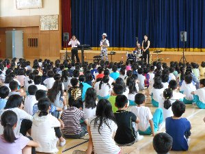 学校演奏会で体育館で演奏する人たちとそれを聞く生徒たちの写真