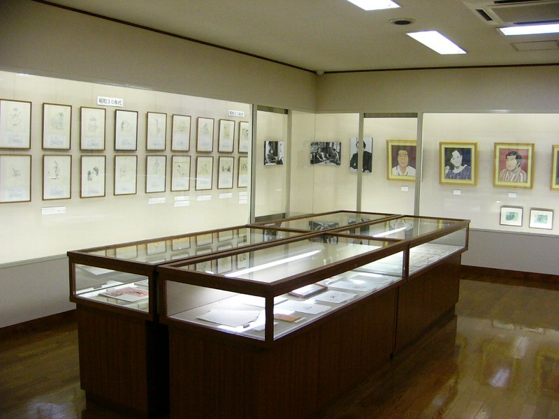 透明の展示ケースが部屋の中央に置かれ、壁際には額縁に入った絵が並べて飾られている写真